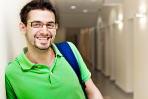Male college student with a backpack and glasses, leaning against a university hallway, smiling at the camera | Transferring to college can be tough with Autism, Aspergers, or other ASD. Blog series about neurodiversity in college for young adults with autism. Can use social skills groups, social skills classes and autism support groups in Palo Alto, CA 94306